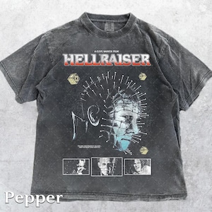May include: Dark grey t-shirt featuring the movie title "Hellraiser" in red and white lettering. The shirt displays a graphic of the character Pinhead with nails in his head. The shirt also has the text "A Clive Barker Film".