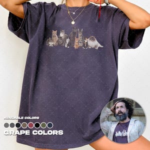 May include: A dark grey, short-sleeved t-shirt featuring a graphic print of nine cats in various colours and breeds arranged in a row across the chest. The shirt has a crew neckline and a relaxed fit. Text at the bottom reads AVAILABLE COLOURS.