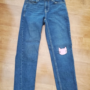 May include: Blue denim jeans with a pink cat face patch on the right knee. The jeans feature a classic five-pocket design and a button and zip closure. The jeans are laid flat on a wooden surface.