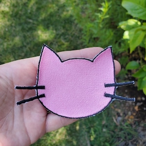 May include: A pink embroidered cat face patch with a black stitched outline and two black whiskers extending from each side. The patch features a simple, flat design with a textured fabric surface, shaped like a feline head with pointed ears.