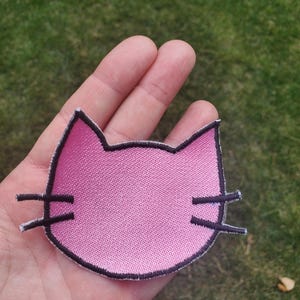 May include: A pink embroidered cat head patch with a black stitched outline and black whiskers on each side. The patch features a textured fabric surface and a distinct cat ear shape, designed for application onto various textile surfaces.