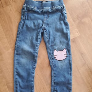 Puede incluir: Pantalones vaqueros azules con cintura elástica y un parche de cara de gato rosa con bigotes bordados en negro en la rodilla izquierda. Los pantalones tienen un diseño clásico de cinco bolsillos con costuras visibles y etiqueta interior.