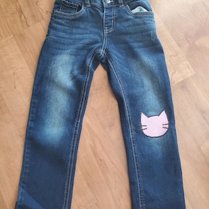 May include: Dark blue denim jeans with a straight leg fit, featuring a pink cat face patch on the left knee. The waistband includes a button closure and the Cat & Jack brand label. The fabric has a faded wash effect on the thighs and front.