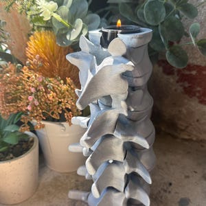 May include: A silver-coloured candle holder shaped like a human spinal column. The vertical structure features stacked vertebrae segments with a cylindrical opening at the top, currently holding a small black tea light candle with a lit flame.