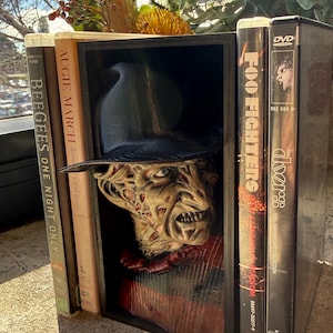 May include: A black 3D-printed book nook insert featuring a sculpted bust of a character with a scarred face, wearing a black fedora and a red and black striped sweater, positioned between a Bee Gees DVD, a book, and two other music DVDs.