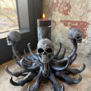 May include: A dark grey candle holder featuring a central pillar with a lit candle, surrounded by multiple sculpted skulls and curved, horn-like protrusions extending outwards in a circular arrangement, finished with a weathered, textured appearance.
