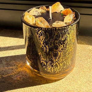 May include: A cylindrical glass candle container featuring a black and gold snakeskin pattern. The top surface is black wax embedded with several translucent, amber-coloured crystals arranged around a single white cotton wick in the centre.