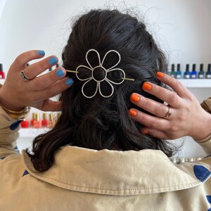 Puede incluir: Una pinza para el pelo de plata en forma de margarita con una barra dorada en el centro. La pinza est&aacute; en el pelo oscuro de una mujer.