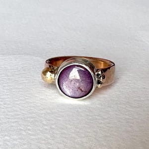 May include: A ring featuring a large, round, purple gemstone set in a silver bezel. The band is a mix of gold and silver, with gold accents. The ring is a statement piece, showcasing a unique blend of colours and textures.