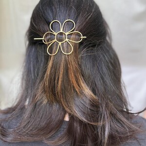 Puede incluir: Una pinza para el pelo de metal dorado en forma de flor con un c&iacute;rculo en el centro. La pinza est&aacute; en el pelo de una persona.