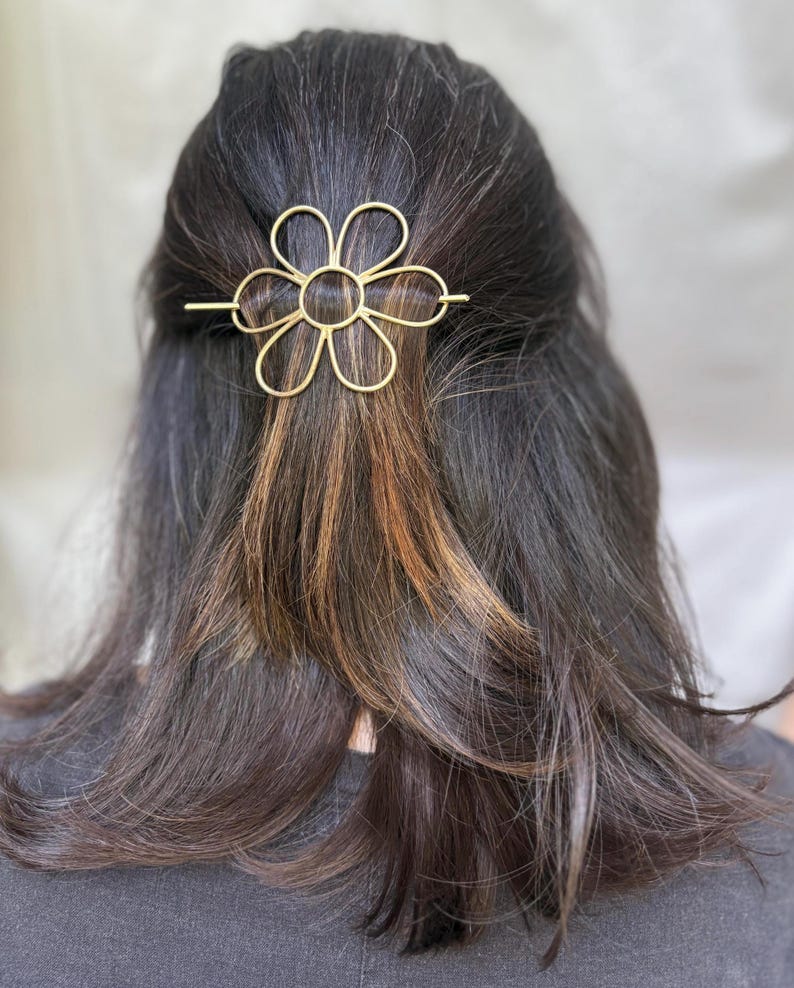 Puede incluir: Una pinza para el pelo de metal dorado en forma de flor con un c&iacute;rculo en el centro. La pinza est&aacute; en el pelo de una persona.