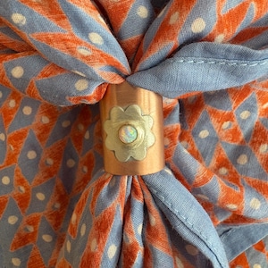 Op de afbeelding: Een blauwe en oranje bandana met een wit stippenpatroon is vastgebonden met een koperen en zilveren ring in de vorm van een bloem.