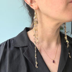 May include: Gold star and pearl chain earrings with a dangling design. The earrings are long and feature a delicate chain with small gold stars and white pearls.
