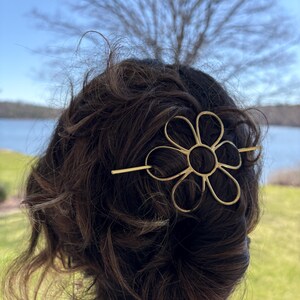 Puede incluir: Una pinza para el pelo de metal dorado en forma de flor con una barra larga y fina que atraviesa la flor. La pinza est&aacute; en el pelo de una persona.