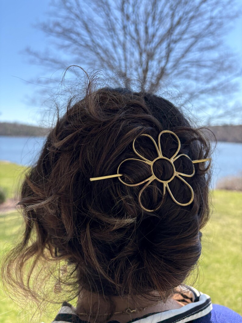 Puede incluir: Una pinza para el pelo de metal dorado en forma de flor con una barra larga y fina que atraviesa la flor. La pinza est&aacute; en el pelo de una persona.