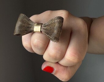 May include: A gold-toned ring featuring a mesh bow design. The ring is worn on a finger with red nail polish.