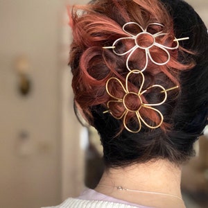Puede incluir: Dos pinzas para el pelo con forma de flor en el pelo de una persona. La pinza de arriba es plateada y la de abajo es dorada.