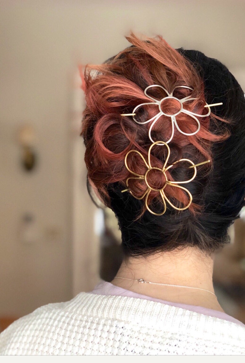 Puede incluir: Dos pinzas para el pelo con forma de flor en el pelo de una persona. La pinza de arriba es plateada y la de abajo es dorada.