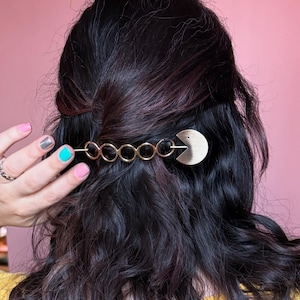 Puede incluir: Una pinza para el pelo dorada con una cadena de círculos unidos y un diseño en forma de Pac-Man. La pinza está en el cabello castaño oscuro de una mujer.