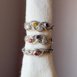 May include: Three silver rings with interwoven bands and colourful gemstones. The rings feature a red, purple, and yellow gemstone.