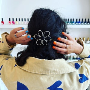 Puede incluir: Una persona con cabello oscuro que lleva una camisa beige con corazones azules impresos en ella. La persona lleva una pinza para el cabello en forma de flor plateada en el cabello. La persona tiene esmalte de u&ntilde;as azul y naranja en las u&ntilde;as.