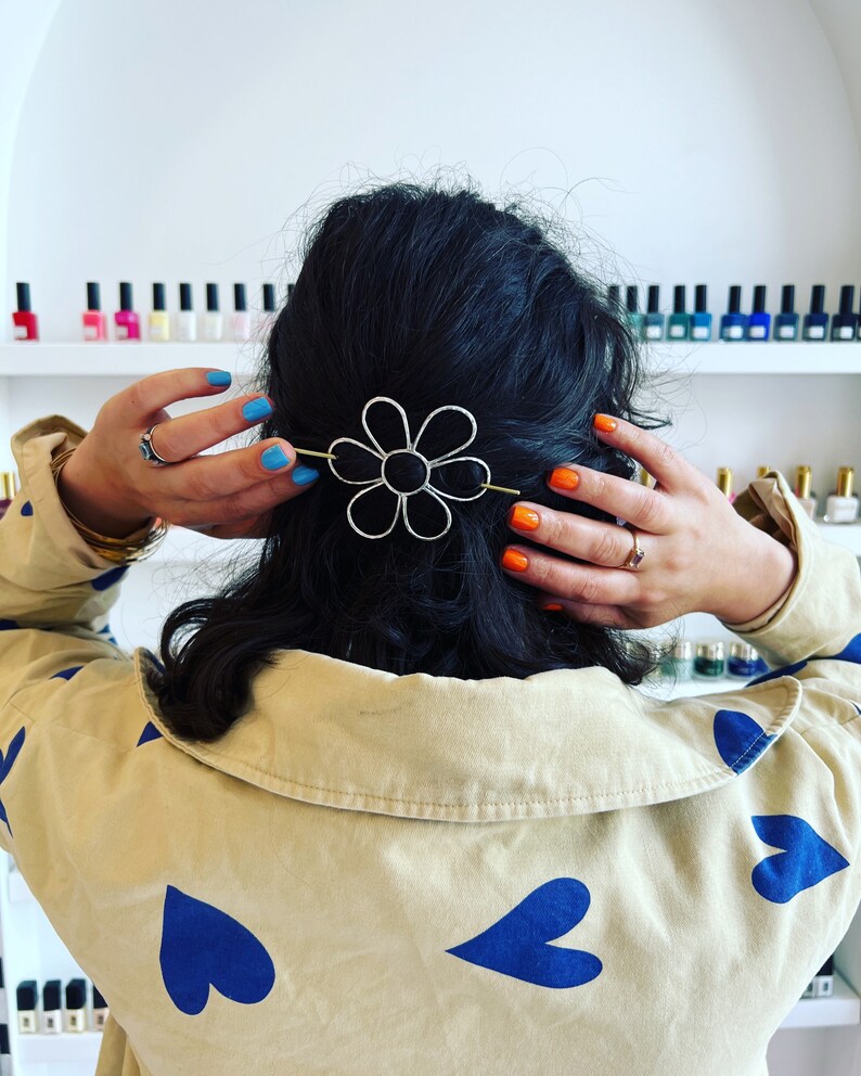 Puede incluir: Una persona con cabello oscuro que lleva una camisa beige con corazones azules impresos en ella. La persona lleva una pinza para el cabello en forma de flor plateada en el cabello. La persona tiene esmalte de u&ntilde;as azul y naranja en las u&ntilde;as.