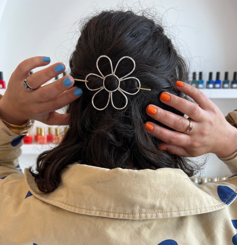 Puede incluir: Una pinza para el pelo de plata en forma de margarita con una barra dorada en el centro. La pinza est&aacute; en el pelo oscuro de una mujer.