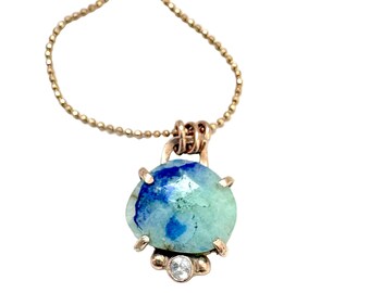 Op de afbeelding: Een goudkleurige ketting met een hanger met een blauwgroene edelsteen. De ovaalvormige edelsteen heeft een facetgeslepen oppervlak. Onder de hoofdsteen is een kleine, heldere edelsteen gezet.