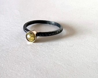 May include: A black ring with a textured band and a gold bezel setting. The ring features a round, light green gemstone. The ring is set against a white background.
