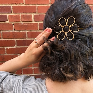 Puede incluir: Una pinza para el pelo de metal dorado en forma de flor con un dise&ntilde;o sencillo. La pinza se lleva en el pelo de una persona.