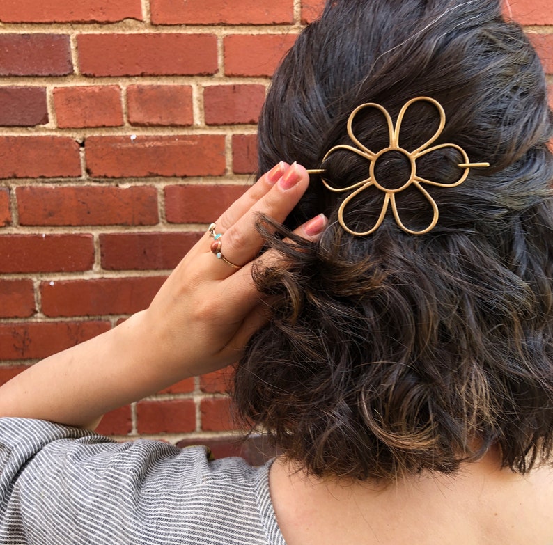 Puede incluir: Una pinza para el pelo de metal dorado en forma de flor con un dise&ntilde;o sencillo. La pinza se lleva en el pelo de una persona.