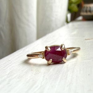 May include: A gold ring featuring a central oval-shaped, faceted deep red ruby gemstone held by four prongs. The band is accented on each side of the central stone by a single small, clear, round-cut white diamond set into the gold metal band.