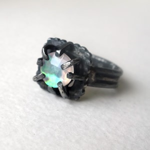 May include: A silver ring featuring a faceted, iridescent gemstone. The stone is set in a dark, textured metal setting with claw-like prongs. The ring band has a ribbed design.