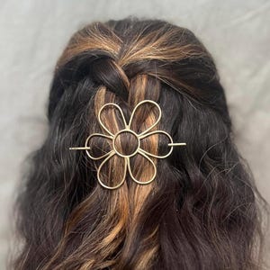 Puede incluir: Una pinza para el pelo de metal dorado en forma de flor con una barra larga y fina que atraviesa el centro de la flor. La pinza est&aacute; en el pelo de una persona.