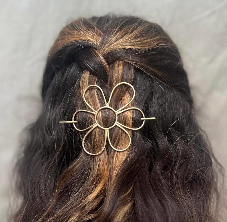 Puede incluir: Una pinza para el pelo de metal dorado en forma de flor con una barra larga y fina que atraviesa el centro de la flor. La pinza est&aacute; en el pelo de una persona.