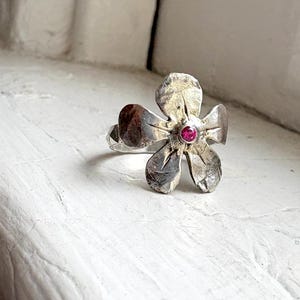 May include: A silver-toned flower ring with a small, round, pink gemstone at its center. The ring's petals have a textured, detailed appearance, and the band is visible. The ring is set against a neutral background.