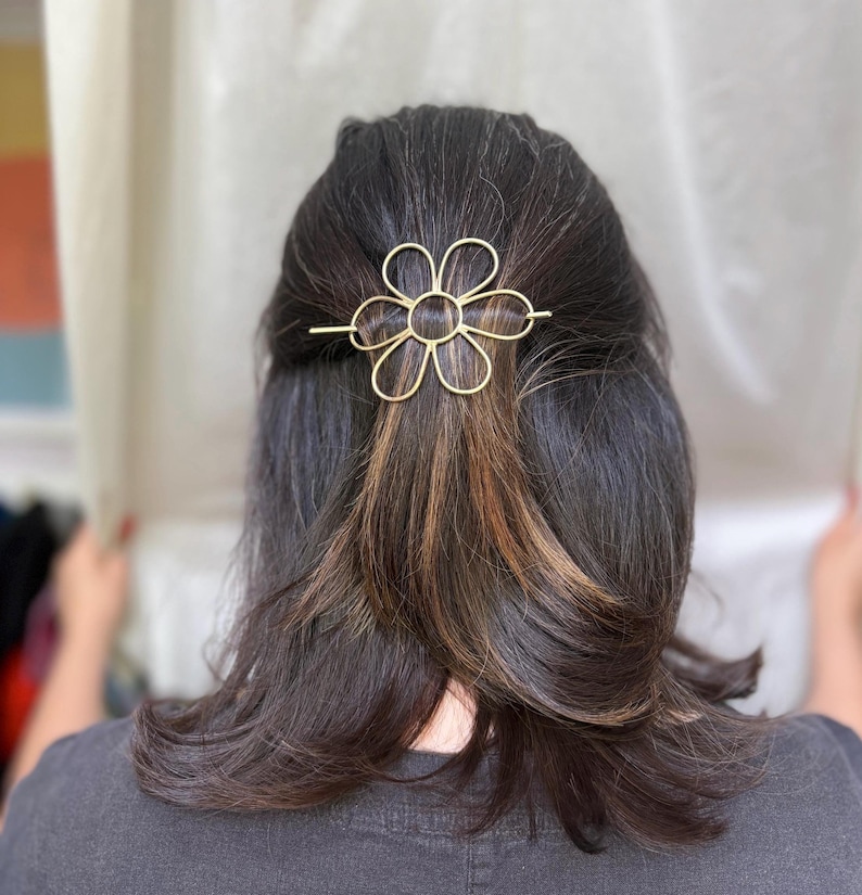 Puede incluir: Una pinza para el pelo de metal dorado en forma de flor con un dise&ntilde;o sencillo. La pinza se utiliza para sujetar el pelo de una persona.
