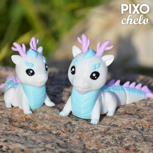 Peut inclure: Deux figurines de dragons blancs avec des accents bleus et lavande. Les dragons ont de grands yeux noirs, des cols bleus et des cornes lavande. Le texte "PIXO chelo" est dans le coin sup&eacute;rieur droit.