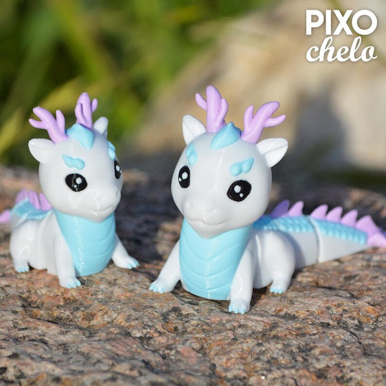 Peut inclure: Deux figurines de dragons blancs avec des accents bleus et lavande. Les dragons ont de grands yeux noirs, des cols bleus et des cornes lavande. Le texte "PIXO chelo" est dans le coin sup&eacute;rieur droit.