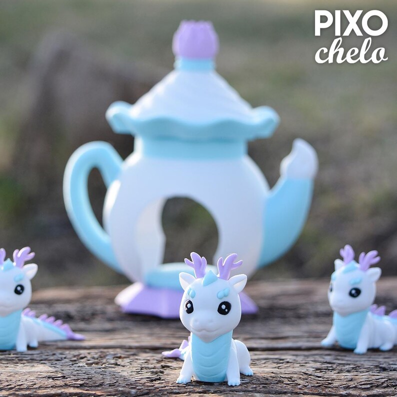 Peut inclure: Une sc&egrave;ne fantaisiste avec une th&eacute;i&egrave;re blanche et bleu clair, surmont&eacute;e d'un couvercle violet, et trois figurines de petits dragons. Les dragons sont blancs avec des accents bleus et des bois violets, cr&eacute;ant un th&egrave;me fantastique.
