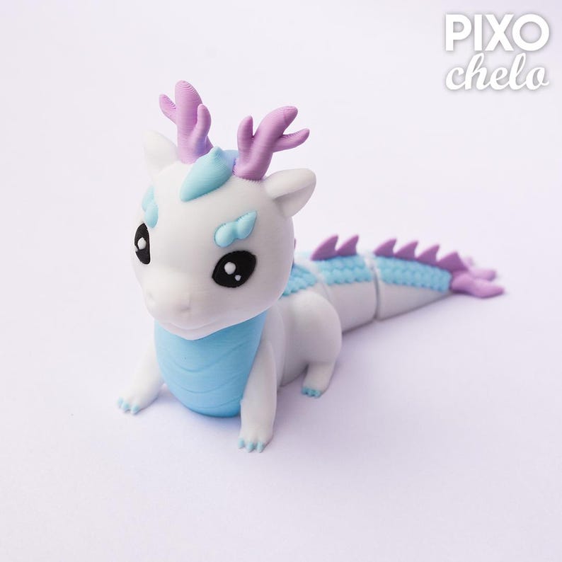 Peut inclure: Figurine de dragon blanc et bleu avec des bois violets et une queue segment&eacute;e. Le dragon a de grands yeux noirs et une poitrine bleu clair. L'image comprend le texte "PIXO chelo" dans le coin sup&eacute;rieur droit.