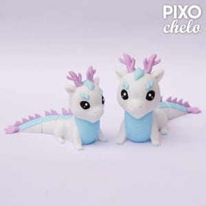 Peut inclure: Deux figurines de dragons blancs avec des d&eacute;tails bleu clair et lavande. Les dragons ont de grands yeux noirs, des bois lavande et des &eacute;pines bleu clair. Le texte "PIXO chelo" est dans le coin sup&eacute;rieur droit.