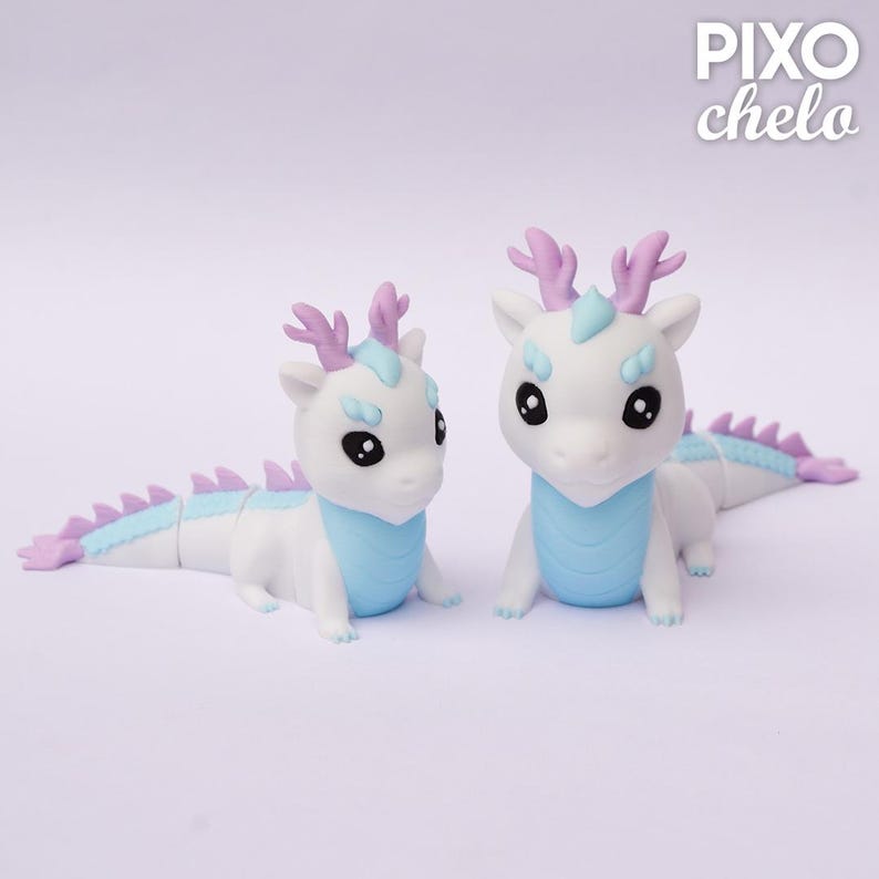 Peut inclure: Deux figurines de dragons blancs avec des d&eacute;tails bleu clair et lavande. Les dragons ont de grands yeux noirs, des bois lavande et des &eacute;pines bleu clair. Le texte "PIXO chelo" est dans le coin sup&eacute;rieur droit.