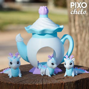 Peut inclure: Une th&eacute;i&egrave;re d&eacute;corative blanche et bleu clair avec un motif floral sur le dessus, accompagn&eacute;e de trois petites figurines de dragons. Les dragons sont blancs et bleu clair avec des accents violets. Le texte "PIXO chelo" est visible.