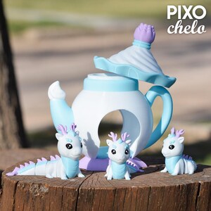 Peut inclure: Une th&eacute;i&egrave;re blanche et bleu clair avec un couvercle assorti, accompagn&eacute;e de trois figurines de dragons. Les dragons ont des d&eacute;tails bleus et des bois violets. Le texte "PIXO chelo" est visible.