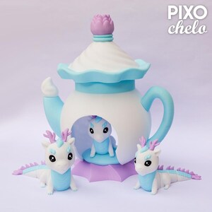 Peut inclure: Une sculpture fantaisiste en forme de th&eacute;i&egrave;re aux couleurs pastel, avec un corps blanc, des accents bleu clair et une base lavande. Trois petites figurines de dragons blancs avec des d&eacute;tails bleus et lavande sont dispos&eacute;es autour de la th&eacute;i&egrave;re. Le texte "PIXO chelo" est visible.