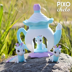 Peut inclure: Une th&eacute;i&egrave;re fantaisiste aux couleurs pastel avec une figurine de dragon &agrave; l'int&eacute;rieur, flanqu&eacute;e de deux figurines de dragon suppl&eacute;mentaires. La th&eacute;i&egrave;re est blanche, bleu clair et lavande, avec un dessus en forme de fleur violette. Le texte "PIXO chelo" est visible.