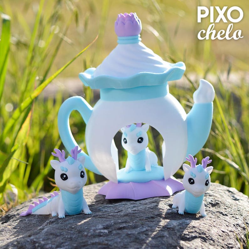 Peut inclure: Une th&eacute;i&egrave;re fantaisiste aux couleurs pastel avec une figurine de dragon &agrave; l'int&eacute;rieur, flanqu&eacute;e de deux figurines de dragon suppl&eacute;mentaires. La th&eacute;i&egrave;re est blanche, bleu clair et lavande, avec un dessus en forme de fleur violette. Le texte "PIXO chelo" est visible.
