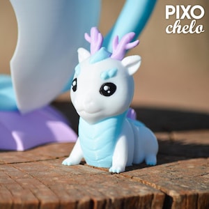 Peut inclure: Figurine de dragon blanc et bleu clair avec des bois violets. Le dragon a de grands yeux noirs et est assis sur une surface en bois. La figurine a une poitrine et un dos bleu clair. Le texte "PIXO chelo" est dans le coin sup&eacute;rieur droit.