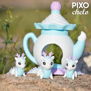 Peut inclure: Une th&eacute;i&egrave;re fantaisiste aux couleurs pastel, avec une anse et des d&eacute;tails bleu clair, et un corps blanc. Trois petites figurines de dragon, blanches avec des d&eacute;tails bleu clair et lavande, sont devant la th&eacute;i&egrave;re. Le texte "PIXO chelo" est visible.